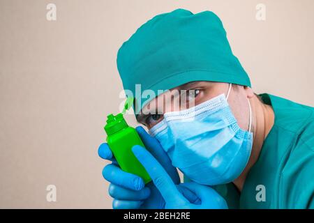 Doktor in grünem Anzug und blauen Maskenhandschuhen mit grüner antiseptischer Flasche in der Hand auf gelbem Hintergrund. Stockfoto