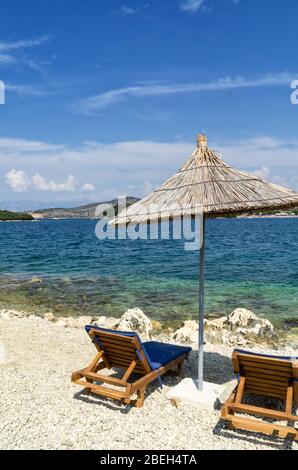 Felsigen Strand am Ufer des Ionischen Meer mit Sonnenliegen und Sonnenschirme Stroh. Blau und tourquse Meer im Hintergrund, niemand. Sommertag. Ksamil, Al Stockfoto