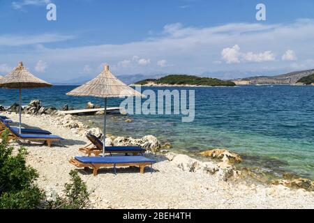 Felsigen Strand am Ufer des Ionischen Meer mit Sonnenliegen und Sonnenschirme Stroh. Blau und tourquse Meer im Hintergrund, niemand. Sommertag. Ksamil, Al Stockfoto