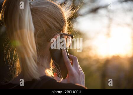 Hinteransicht von hinten, auf der junge Frau im Park bei Sonnenuntergang im Freien mit dem Handy spricht. Mädchen mit Handy, mit digitalem Gerät, Blick auf die Einstellung Stockfoto