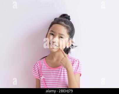 Porträt eines kleinen asiatischen Mädchens mit einem rosa-weiß gestreiften Kleid. Das Kind mit schwarzen Haaren lächelt und zeigt auf ihr Gesicht. Stockfoto