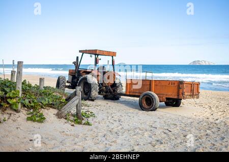 Oranger alter Traktor mit Anhänger am Strand von Ipanema, der für Wartungsarbeiten am leeren Strand mit Meer und Insel im Hintergrund geparkt wurde Stockfoto