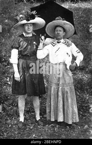Zwei Männer, beide bekleideten sich Kopf bis Fuß in Frauenkleidung (außer ihren Schuhen), um 1906. Für die Edwardian Ära, beide sind fein gekleidet in lässige Sommerzeit Kleidung, einschließlich enorm breit-randvoll, geblümten Hüte. Der Mann, der den offenen Schirm hielt (im Schatten stehend), hatte ein zierliches Taschentuch an seine weiße Bluse geklemmt. Sind sie ein liebevolles Paar? Freunde? Schwul? Vielleicht sind sie für ein Theaterstück in Kostüme gekleidet. Um alle meine verwandten Bilder zu sehen Suche: Prestor Vintage Gay Stockfoto