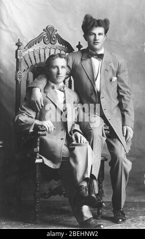 Zwei Männer teilen sich einen fein geschnitzten Holzstuhl in einem Fotostudio um 1914. Sie sind offensichtlich nah beieinander und sehr schön gekleidet. Beachten Sie die gestreiften Socken! Sie tragen Fliege, Westen, scharfe Anzüge, haben Taschentücher in Jackentaschen, und ihre Hose ist richtig geknittert. Irgendwie erinnert mich die Frisur des Mannes auf der rechten Seite an den Entertainer des 20. Jahrhunderts, Liberace. Sind sie ein liebevolles Paar, Brüder? Freunde? Schwul? Um alle meine verwandten Bilder zu sehen Suche: Prestor Vintage Gay Stockfoto