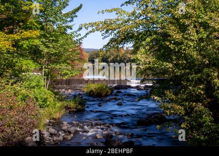 Wasser rauscht über einem Damm vor dem Hintergrund der früherbstlichen Blätter in Vermont, Stockfoto