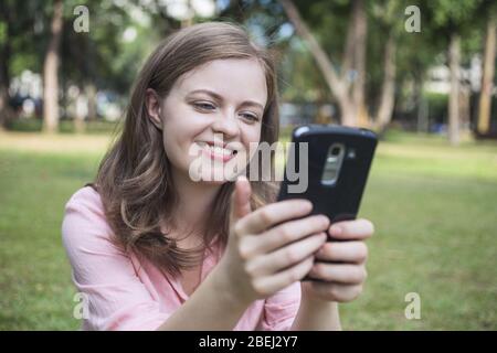 Die junge kaukasische Frau, die lächelnd auf den Bildschirm des Telefons schaut, sieht glücklich aus. Außenbilder. Stockfoto