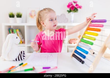 Kleines lächelndes blondes Mädchen, das am weißen Schreibtisch sitzt und auf den bunten Abakus im Klassenzimmer zählt. Vorschulbildung Stockfoto