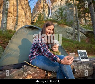 Frau und Kind machen ein Selfie auf einem Handy auf dem Hintergrund eines touristischen Zeltes mit einem Solarpanel am Fuße einer Klippe im Wald. Familie in den Sonnenstrahlen mit sanftem Lächeln Stockfoto