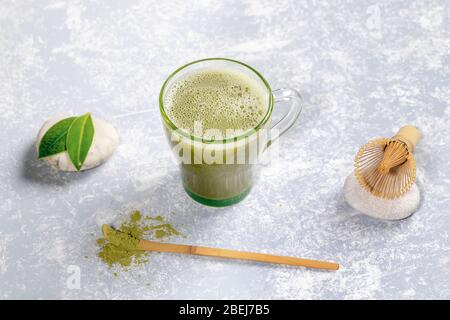 Grüner Glasbecher mit japanischem Matcha-Grüntee, spezieller Holzlöffel mit Pulver, Bambusbesen zum Peitschen und Blätter auf Steinen auf grauem texturiertem Boden Stockfoto