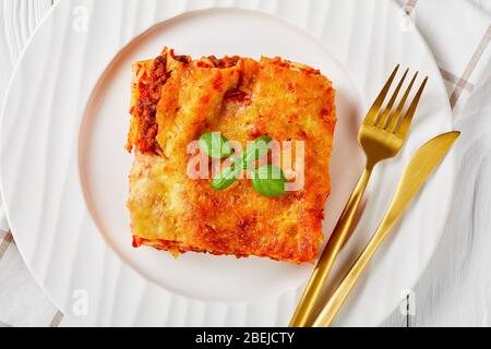 Fleisch Cannelloni mit Tomatensauce und geschmolzenem Mozzarella Käse und frischem Basilikum auf einem weißen Teller mit Besteck auf weißem Holzhintergrund gebacken, Stockfoto
