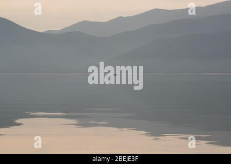 Fischerboot in der Ferne vor der Kulisse einer schönen Bergkette mit einer malerischen Spiegelung im See. Hintergrund in minimalistischer Stockfoto