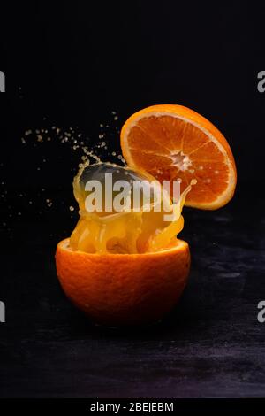 Nahaufnahme eines frischen Orangensaftes, der von einer in zwei Hälften geschnittenen Orange auf einem dunklen Hintergrund gespritzt wird. Stockfoto