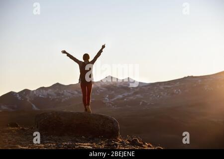 Glückliche Wanderin mit Rucksack steht mit erhobenen Armen auf großem Felsen und blickt auf Berge und Sonnenuntergang Stockfoto