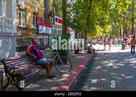 Kerch, Russland - 13. August 2019: Ein Mann sitzt auf einer Bank und hört einem Straßenmusiker zu und Fußgänger Touristen gehen auf der Straße von Th Stockfoto