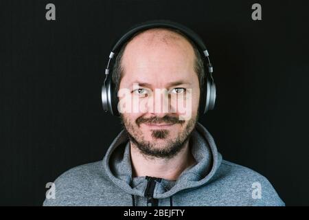Bärtiger entspannter Mann im Hoodie, der mit Kopfhörern vor schwarzem Hintergrund steht, Musik hört, Kopf und Schultern Stockfoto