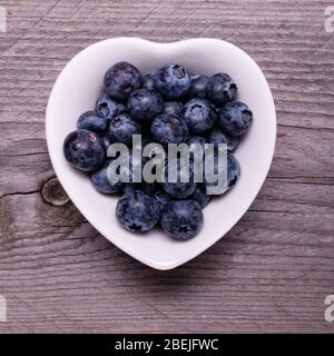 In der weißen keramischen herzförmigen Schale, reife Heidelbeeren, im Vordergrund, vor dem Hintergrund aus rauem Holz Stockfoto