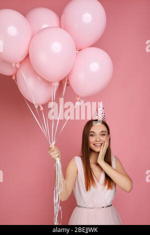 Schöne, überraschte glückliche Frau im Kleid und Geburtstagshut, die mit Pastellpinken Luftballons auf pinkfarbenem Hintergrund feiert. Süßes glückliches junges Mädchen auf einer Geburtstagsparty. Stockfoto