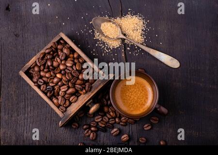Auf dem rohen und dunklen Holztisch eine Tasse Kaffee mit einem Löffel braunem Zucker und gerösteten Kaffeebohnen. Stockfoto