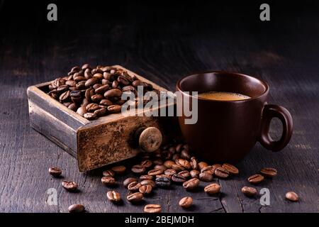 Auf dem rohen und dunklen Holztisch eine Tasse heißen Kaffee und eine Schublade einer alten Mühle mit gerösteten Kaffeebohnen. Stillleben Stockfoto