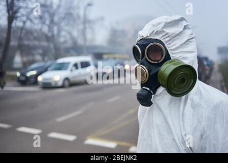 Selektiver Fokus des Wissenschaftlers trägt Gasmaske und Schutzkostüm Blick auf Straße mit Autos, warten auf grünes Licht. Nahaufnahme eines Mannes, der sich in der Nähe einer Fußgängerüberfahrt befindet. Umweltkatastrophe, Nebel. Stockfoto