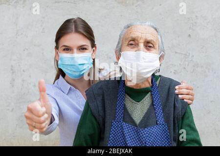 Porträt einer freundlichen Pflegekraft posiert mit älteren kranken Frau trägt chirurgische Maske wegen Covid-19 Pandemie, zeigt Daumen nach oben Stockfoto