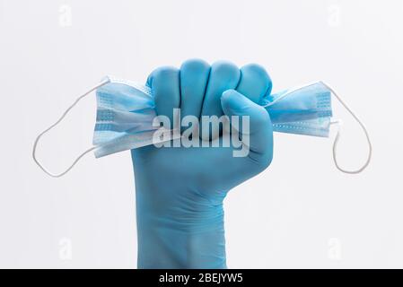 Fausthand in medizinischem blauen Latex-Schutzhandschuh, der eine medizinische Maske als Zeichen der Pandemieresistenz hält - auf weißem Hintergrund. Krankheitszeichen stoppen Stockfoto