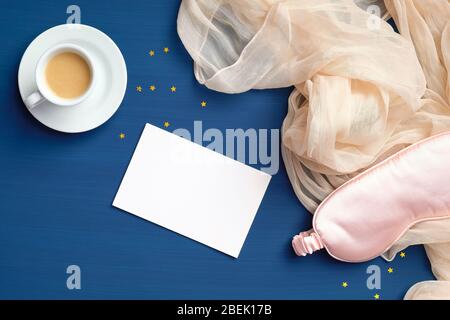 Happy Morning Concept. Flache Lay Zusammensetzung mit Tasse Kaffee, Schlaf Augenmaske, Decke und leere Papierkarte auf dunkelblauem Hintergrund Stockfoto