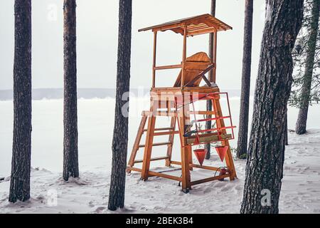 Rettungsschwimmer Turm an einem verschneiten Strand am Fluss. Rettungsschwimmer Turm zwischen Pinien am Ufer eines gefrorenen Flusses Stockfoto