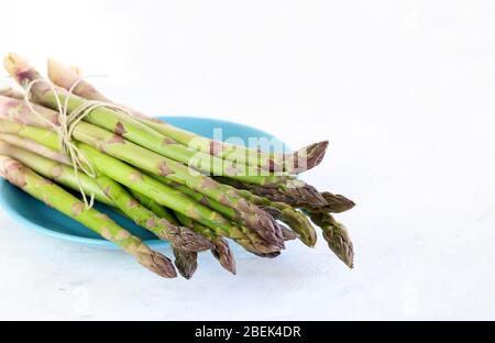 Bündel von grünem frischen Spargel in einem blauen Teller auf weißem Hintergrund. Konzept der gesunden Ernährung. Frisches Gemüse. Nahaufnahme, Kopierbereich. Stockfoto