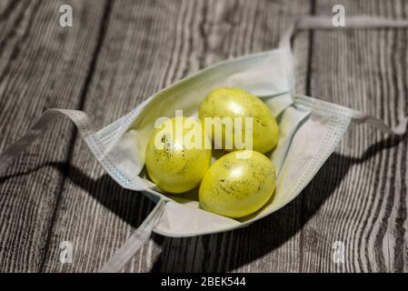 Drei farbige gelbe ostereier, die in medizinischer Maske auf dem Holzhintergrund liegen. Coronavirus Covid-19. Konzeptfoto. Ostern 2020. Stockfoto