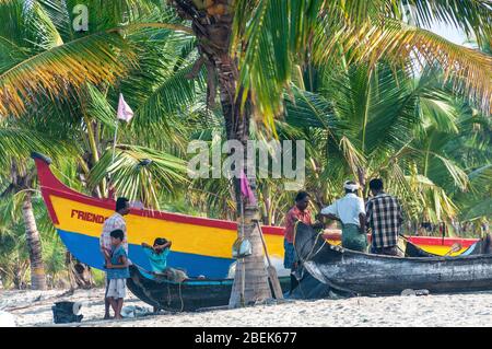 Familien entspannen neben ihren hölzernen Fischerbooten im kleinen Fischerdorf am Marari Beach, Mararikulam, Alapuzha District, Kerala, Indien Stockfoto