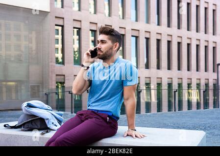 Porträt von fröhlichen modernen Geschäftsmann sprechen per Telefon und lächeln, während im Freien auf einer Bank sitzen Stockfoto