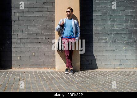 Junger bärtiger Mann, Modelaufstand, im urbanen Hintergrund, der lässige Kleidung trägt und sich an einer Bürowand lehnt Stockfoto