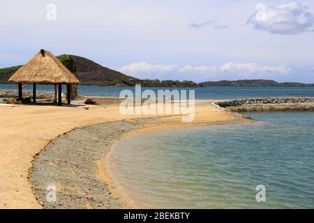 Tiki-Einzelhütte mit Schutz vor der heißen Sonne auf der Insel im Südpazifik, am Rande des warmen Wassers mit Bergkette in der Ferne. Stockfoto