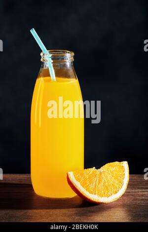 Orangensaft in einer Glasflasche mit Stroh und einer Orangenscheibe auf einem Holztisch mit schwarzem Hintergrund Stockfoto