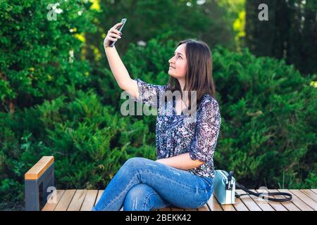 Mädchen mit Telefon auf Bank im Park sitzen. Brunette Frau mit Handtasche Stockfoto