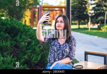 Mädchen mit Telefon auf Bank im Park sitzen. Brunette Frau mit Handtasche Stockfoto