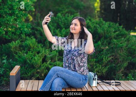 Mädchen mit Telefon auf Bank im Park sitzen. Brunette Frau mit Handtasche Stockfoto