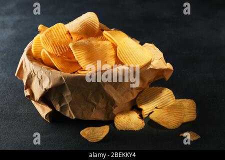 Knusprige Kartoffelchips in einer Schüssel mit Papiertüte auf schwarzem Hintergrund. Stockfoto