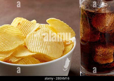 Kartoffelchips in einer weißen Schüssel und ein Glas Cola mit Eis. Makroansicht in der Nähe Stockfoto