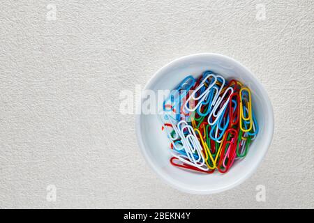 Haufen von bunten Büroklammern in einer weißen Schüssel auf weißem texturiertem Hintergrund.. Makroansicht mit flacher Lay-Ansicht. Stockfoto