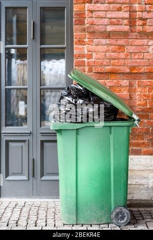 Offener großer grüner Plastikmüllbehälter mit vollen Mülltüten. Entsorgung von Abfällen. Überfüllter Mülltonnen über Ziegelwänden und Türen. Stockfoto