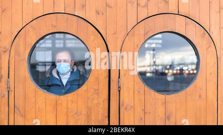 Der Mensch schaut aus dem Fenster, trägt eine medizinische Schutzmaske gegen übertragbare Infektionskrankheiten und als Schutz gegen Grippe, Coronavirus, Covid-19 Stockfoto