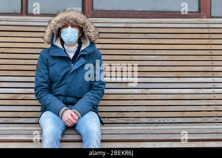 Kranker Mann mit Kapuze, der allein auf der Bank sitzt, Gesichtsmaske gegen übertragbare Infektionskrankheiten trägt, Covid-19. Leere Stadt von Menschen wegen coro Stockfoto