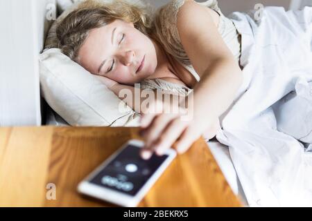 Junge Osteuropäerin, die im Bett liegt und morgens den Wecker auf ihrem Handy abschaltet. Stockfoto