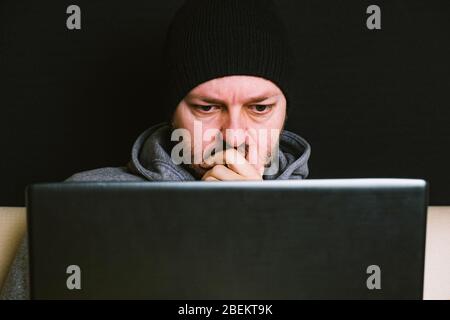 Mann trägt schwarze Mütze und Hoodie sitzt hinter dem Computer-Monitor in der Dunkelheit, Nahaufnahme, hacking Computer-System-Konzept Stockfoto