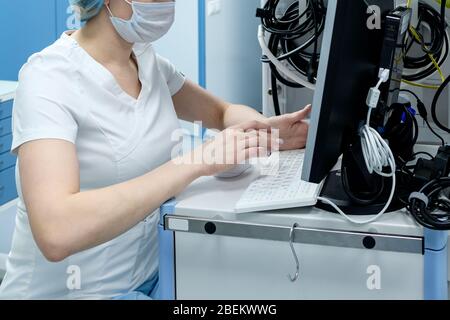 Ein Arzt in einer Maske und Handschuhe arbeitet an einem Computer mit medizinischen Geräten in der Notaufnahme. Analyse von Informationen über Coronavirus infectio Stockfoto