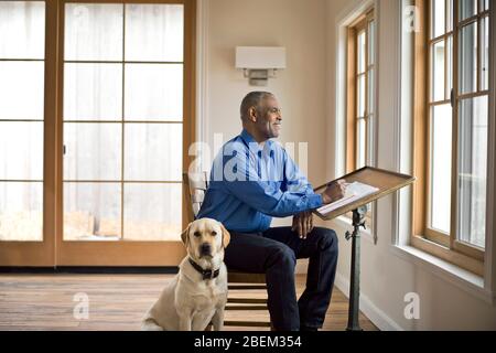 Lächelnder älterer Mann, der an seinem Schreibtisch sitzt und aus dem Fenster mit seinem Hund an seiner Seite blickt Stockfoto