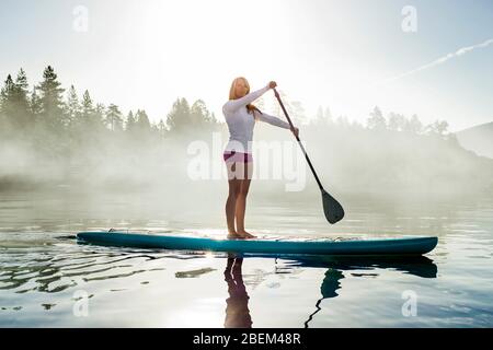 Aktive junge Frau paddeln im Morgennebel über einen See Stockfoto