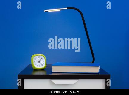 Eine moderne Lampe, ein Wecker und ein Buch auf einem Nachttisch gegen eine blaue Innenwand Stockfoto